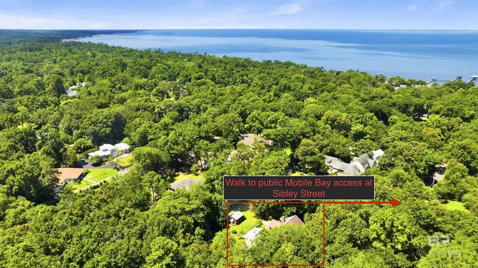 Aerial view of the wooded parcel at 7182 Jewett St in Fairhope, Alabama, with a boxed outline drawn around the cabin's three-lot lot and a Mobile Bay shoreline visible in the upper right of the frame under a clear blue sky