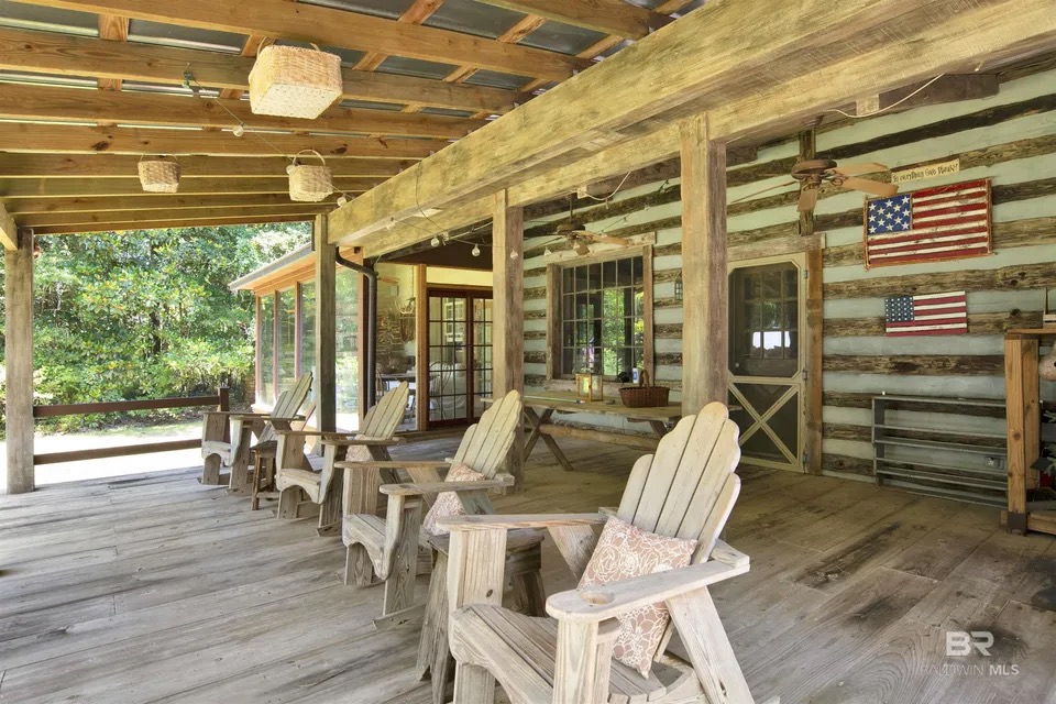 Covered porch at 7182 Jewett St showing exposed rough-hewn cedar log beams and rafters, weathered wide-plank flooring, a row of Adirondack chairs with a small side table, hanging baskets from the rafters, and a small American flag mounted on the log wall