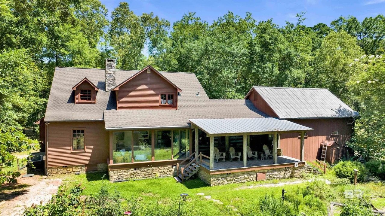 Rear elevation of the cabin at 7182 Jewett St in Fairhope, Alabama, cedar exterior with a stone chimney and dormers, a wraparound covered porch on a stacked-stone base, and a metal-roofed guest wing on the right, framed by mature pines and hardwoods